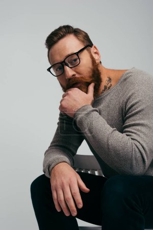 Foto de Retrato de hombre barbudo en gafas tocando la barbilla mientras está sentado en una silla aislada en gris - Imagen libre de derechos