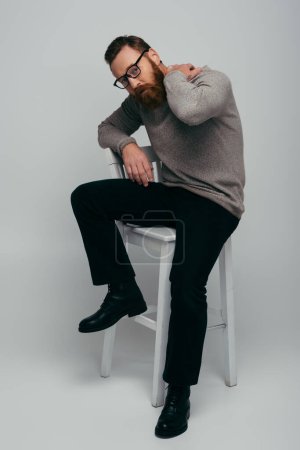 Full length of fashionable bearded man touching neck while sitting on chair on grey background 