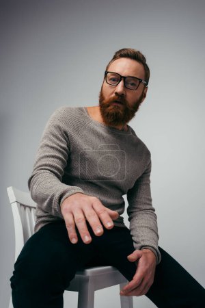 Modischer Mann mit Brille und Pullover blickt in die Kamera, während er isoliert auf einem Stuhl in grau sitzt 