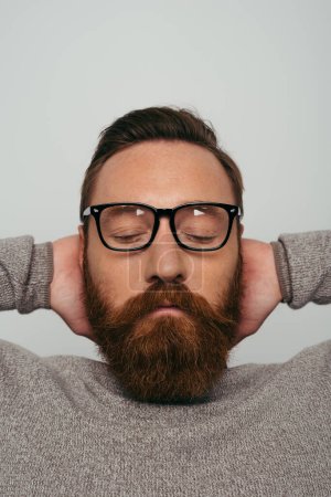 Portrait of bearded man in eyeglasses touching back of head isolated on grey 