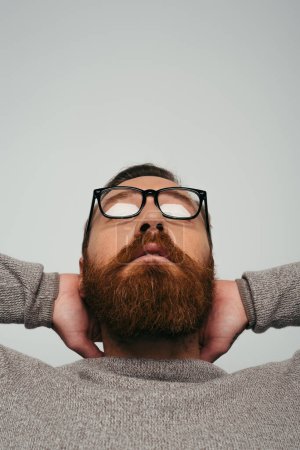 Photo for Bearded man in long sleeve and eyeglasses touching neck isolated on grey - Royalty Free Image
