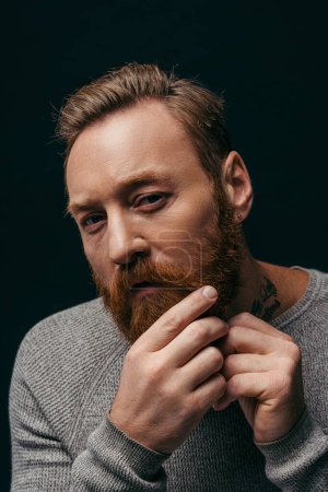 Téléchargez les photos : Portrait d'homme tatoué élégant touchant la barbe et regardant la caméra isolée sur noir - en image libre de droit