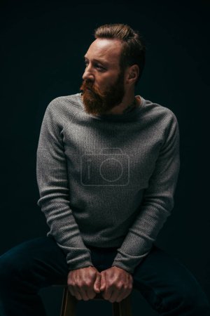 Photo for Fashionable man in sweater looking away while sitting on chair isolated on black with shadow - Royalty Free Image