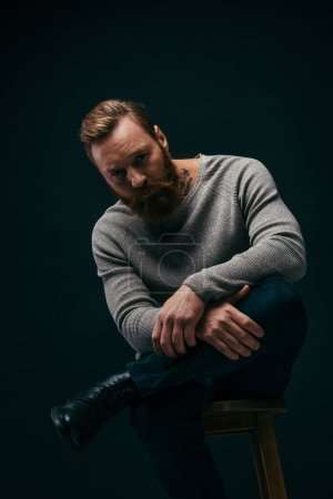 Photo for Tattooed and bearded model in jumper posing on chair isolated on black - Royalty Free Image