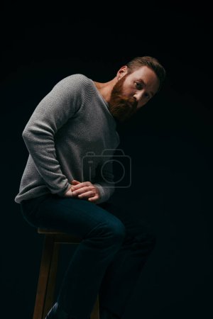 Hombre de moda en jersey posando en silla aislada en negro con sombra 