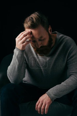 Photo for Bearded man in jumper touching forehead while standing near armchair isolated on black - Royalty Free Image