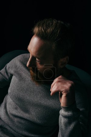 Bärtiger Mann im Pullover schaut weg, während er auf einem Sessel sitzt, isoliert auf schwarz 