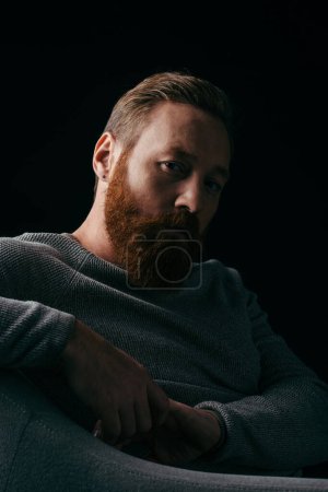 Photo for Stylish bearded man in jumper sitting on armchair isolated on black with shadow - Royalty Free Image