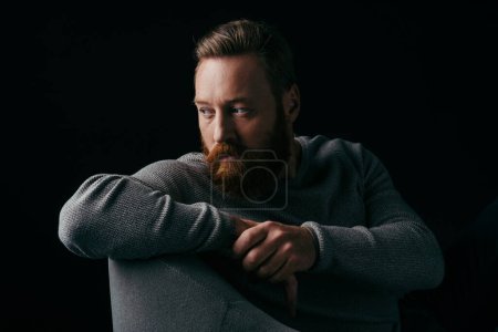Photo for Bearded model in grey jumper looking away while sitting on armchair isolated on black - Royalty Free Image