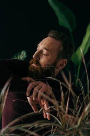 Fashionable man in jumper looking away near blurred plants isolated on black 