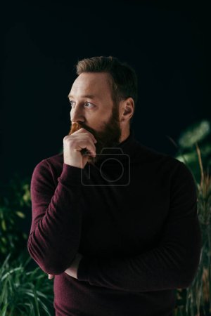 Foto de Elegante hombre en jersey borgoña tocando bigote cerca de plantas aisladas en negro - Imagen libre de derechos