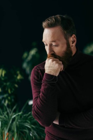 Photo for Pensive bearded man in jumper looking away near blurred plants isolated on black - Royalty Free Image