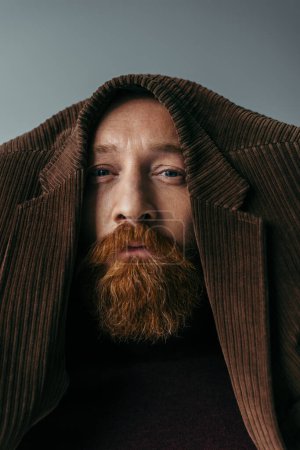 Foto de Hombre barbudo con chaqueta marrón en la cabeza mirando a la cámara aislada en gris - Imagen libre de derechos