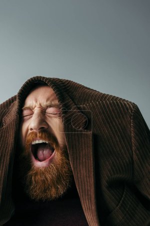 bearded man with closed eyes and brown blazer on head screaming isolated on grey 