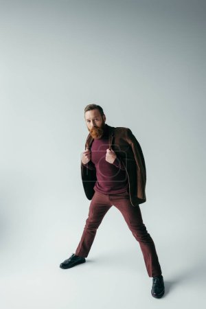 full length of stylish man in blazer and burgundy color outfit posing on grey
