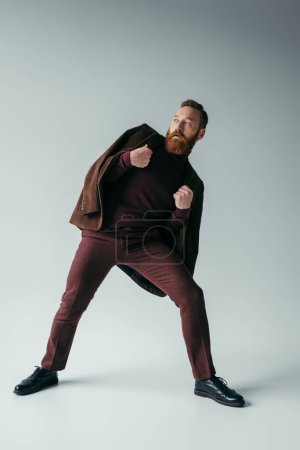 Foto de Longitud completa de hombre con estilo en chaqueta y traje de color burdeos mirando hacia otro lado en gris - Imagen libre de derechos