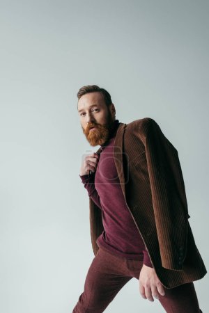 bearded man in turtleneck and brown blazer looking at camera isolated on grey 