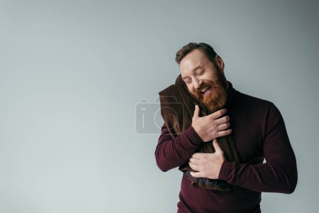 Téléchargez les photos : Homme barbu à col roulé embrassant blazer et souriant isolé sur gris - en image libre de droit