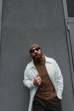 stylish man in beanie hat and sunglasses posing with hand in pocket outside