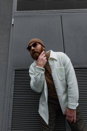 stylish man in beanie hat and sunglasses standing in white shirt jacket and scratching beard outside 