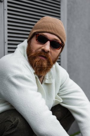 bearded man in stylish sunglasses and beanie hat looking away outdoors