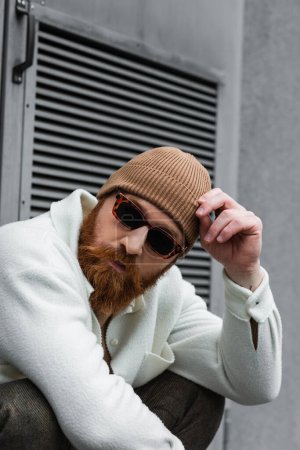 bearded man in stylish sunglasses and trendy beanie hat looking away outdoors
