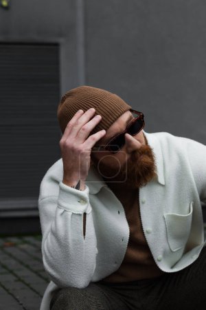 bearded man in stylish sunglasses and white shirt jacket adjusting beanie hat