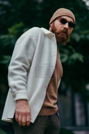 Photo for Bearded man in stylish sunglasses and beanie hat standing in white shirt jacket near green leaves - Royalty Free Image