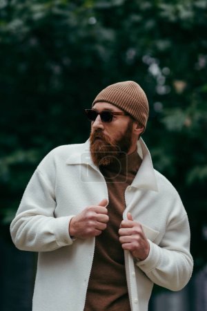 Photo for Bearded man in stylish sunglasses and beanie hat adjusting white shirt jacket near green leaves - Royalty Free Image