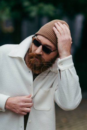 bearded man in stylish sunglasses and white shirt jacket adjusting beanie hat near green leaves