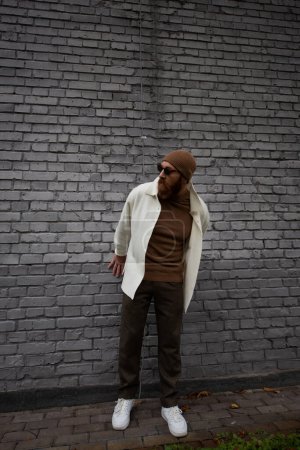 full length of stylish and bearded man in beanie hat and sunglasses standing near brick wall 