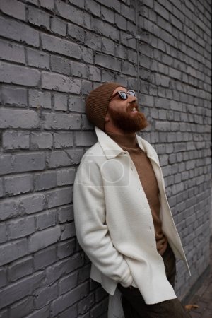 cheerful and bearded man in beanie hat and sunglasses leaning on brick wall 