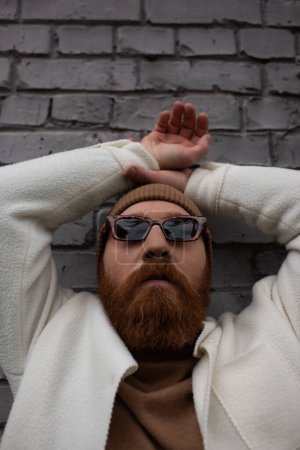 stylish and bearded man in beanie hat and sunglasses near brick wall 