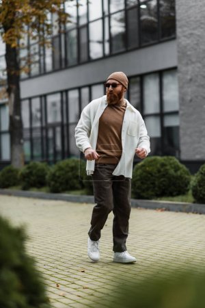 full length of bearded man in beanie hat and stylish sunglasses walking in shirt jacket near modern building 