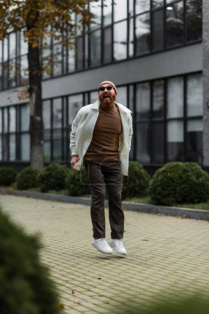 Téléchargez les photos : Homme barbu en bonnet et lunettes de soleil tendance sautant tout en souriant près du bâtiment moderne - en image libre de droit