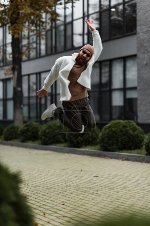 Photo for Full length of excited man in trendy sunglasses and beanie hat jumping near modern building - Royalty Free Image