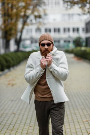hombre barbudo en gorro sombrero y gafas de sol de moda sensación de frío mientras camina fuera