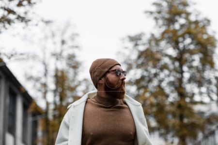 stylish bearded man in beanie hat and trendy sunglasses looking away against trees and cloudy sky 