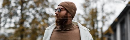 stylish bearded man in beanie hat and trendy sunglasses looking away against trees and cloudy sky, banner