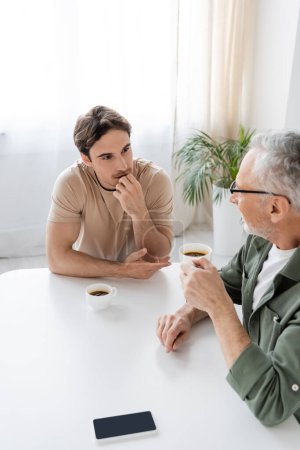 young man holding hand near face while listening to grey haired father near coffee cups in kitchen