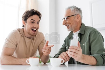 Erstaunter Mann zeigt auf Handy in der Hand von lächelndem Papa, während er in der Küche neben Kaffeetassen sitzt