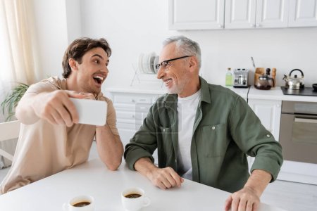 young excited man with mobile phone taking selfie with smiling father near coffee cups in kitchen