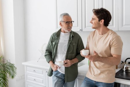 lächelnder älterer Mann mit Kaffeetasse, während er mit der Hand in der Tasche steht und dem Sohn in der Küche zuhört