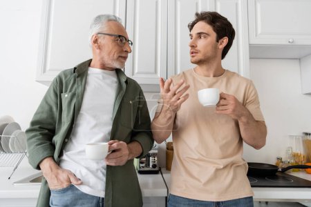Junger Mann mit Kaffeetasse gestikuliert, während er mit Papa spricht, der mit der Hand in der Tasche in der Küche steht