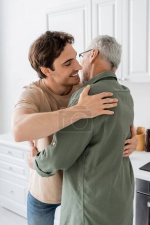 Téléchargez les photos : Jeune homme joyeux embrassant père aux cheveux gris à la maison dans la cuisine - en image libre de droit