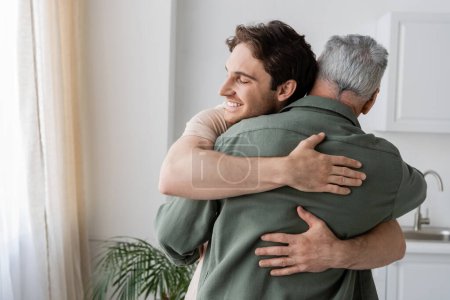 happy young man with closed eyes embracing mature dad in kitchen