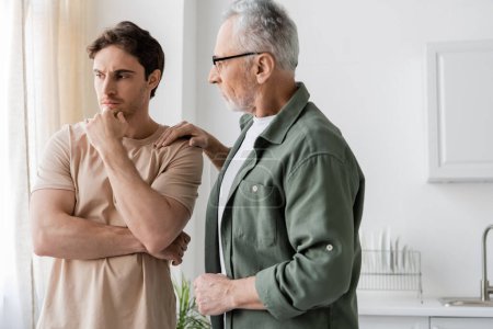 thoughtful and upset man holding hand near face while father calming him in kitchen