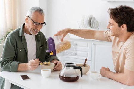 junger Mann gießt Cornflakes in Schüssel neben glücklichem Vater und Kaffeekanne in Küche