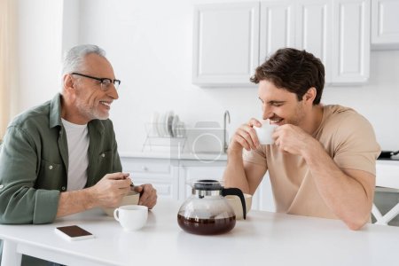Lächelnder Mann schaut Sohn beim Frühstück mit Kaffeetasse an und lacht mit geschlossenen Augen