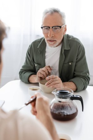 attentive man in eyeglasses looking at blurred son near breakfast and coffee pot on kitchen table